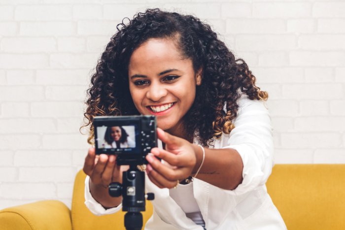 african american woman in white jacket seated on a  yellow sofa adjusts a video camera on a tripod