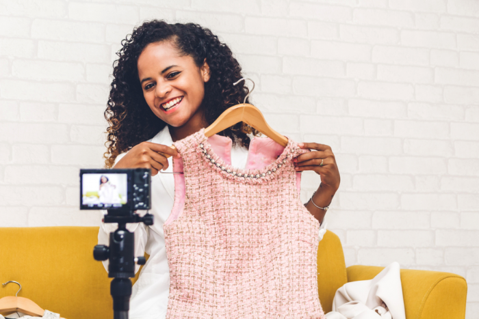 african american woman in a white jacket seated on a yellow couch holds up pink wool houndstooth print dress in front of her to camera and smiles